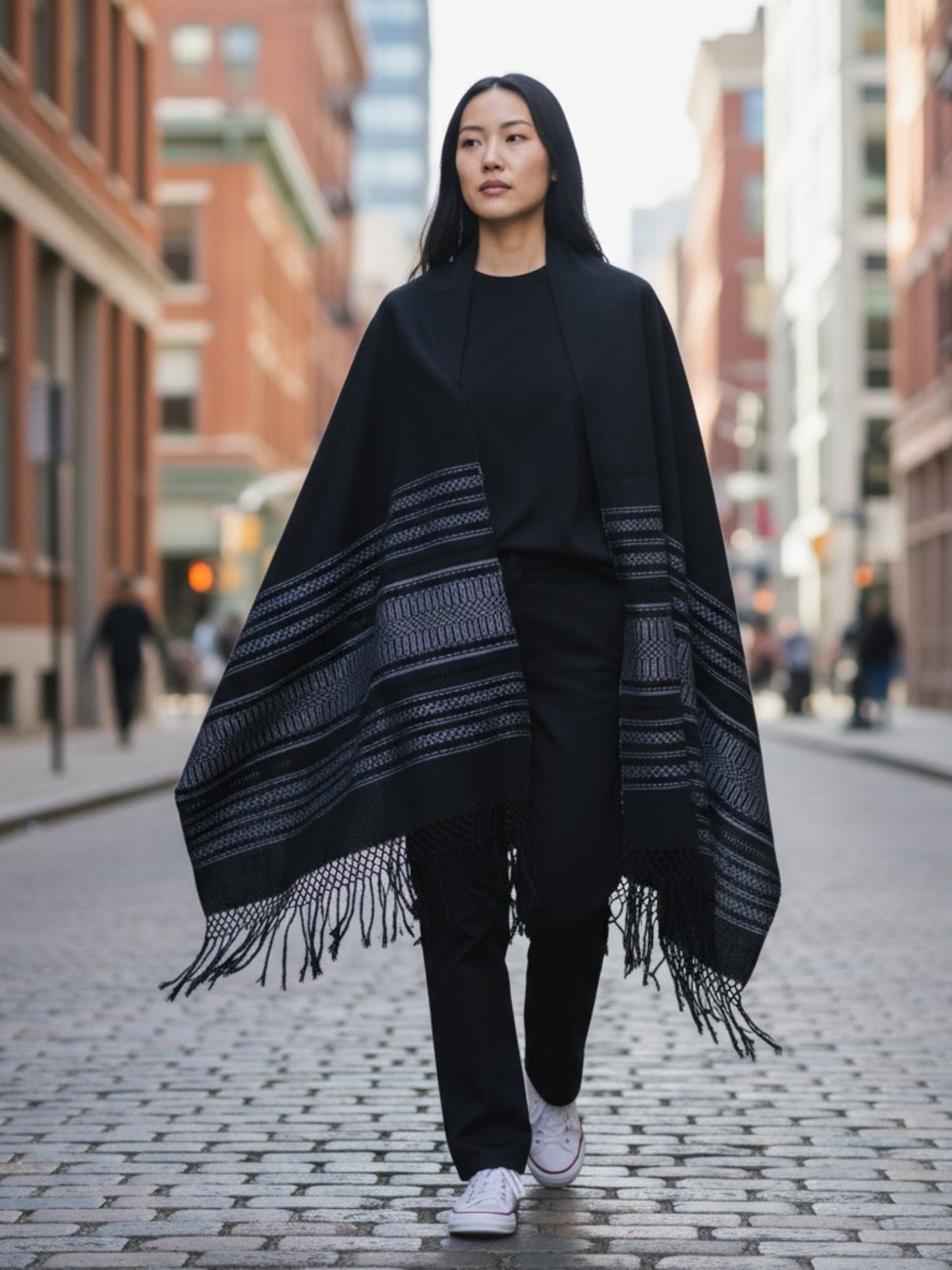 Woman wearing a black handwoven Mexican rebozo shawl with woven borders and fringe, styled as a wrap in an urban street setting