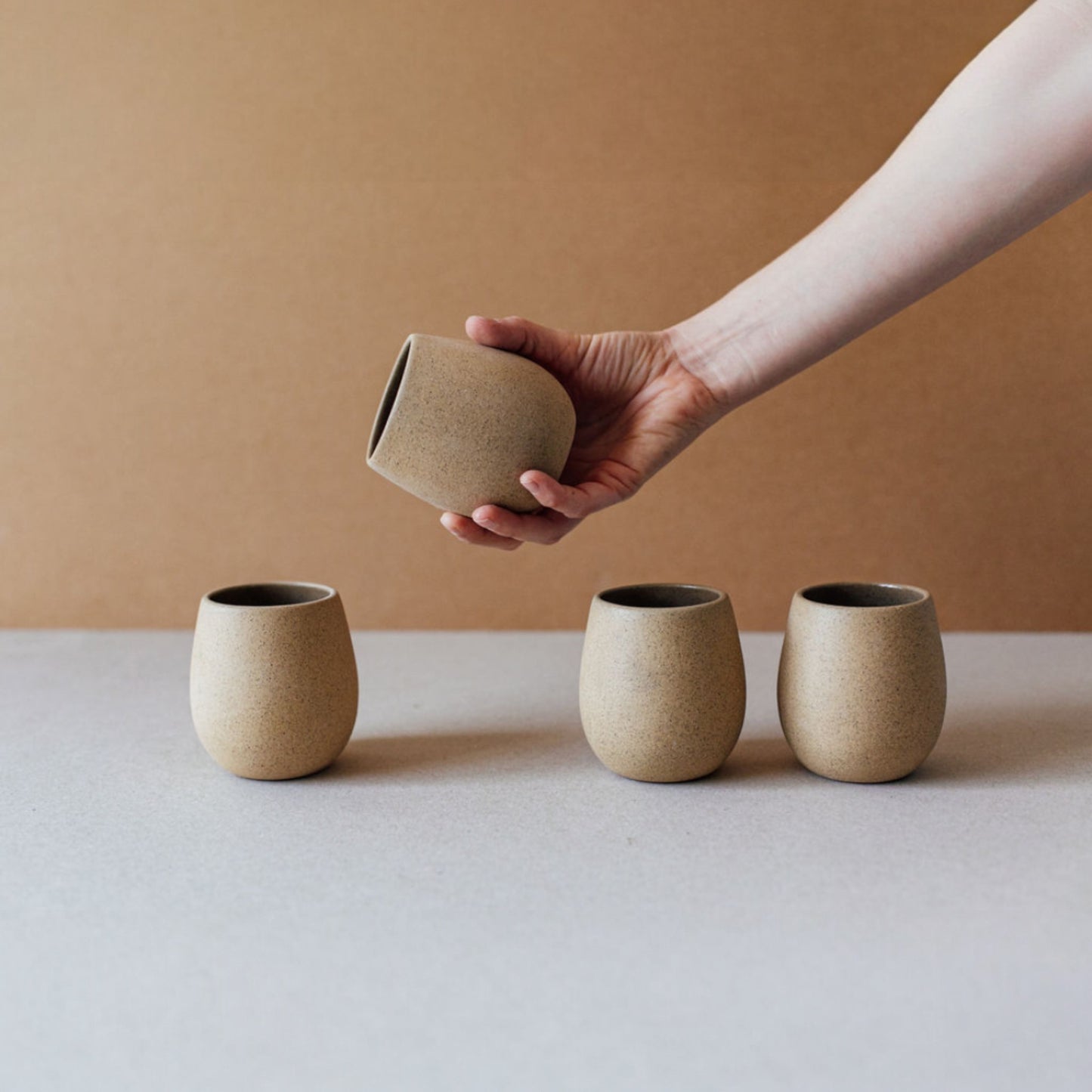 Several round stoneware cups in sand colors arranged in a row showing their sculptural shape.