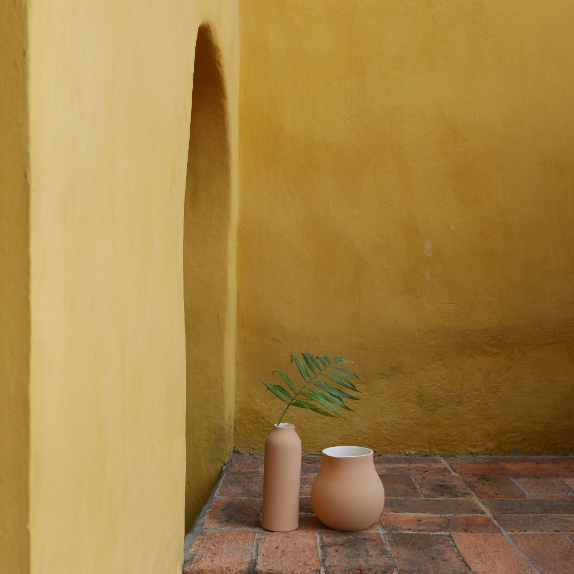 Tall terracotta urn vase placed on rustic terracotta tiles against a yellow textured wall.