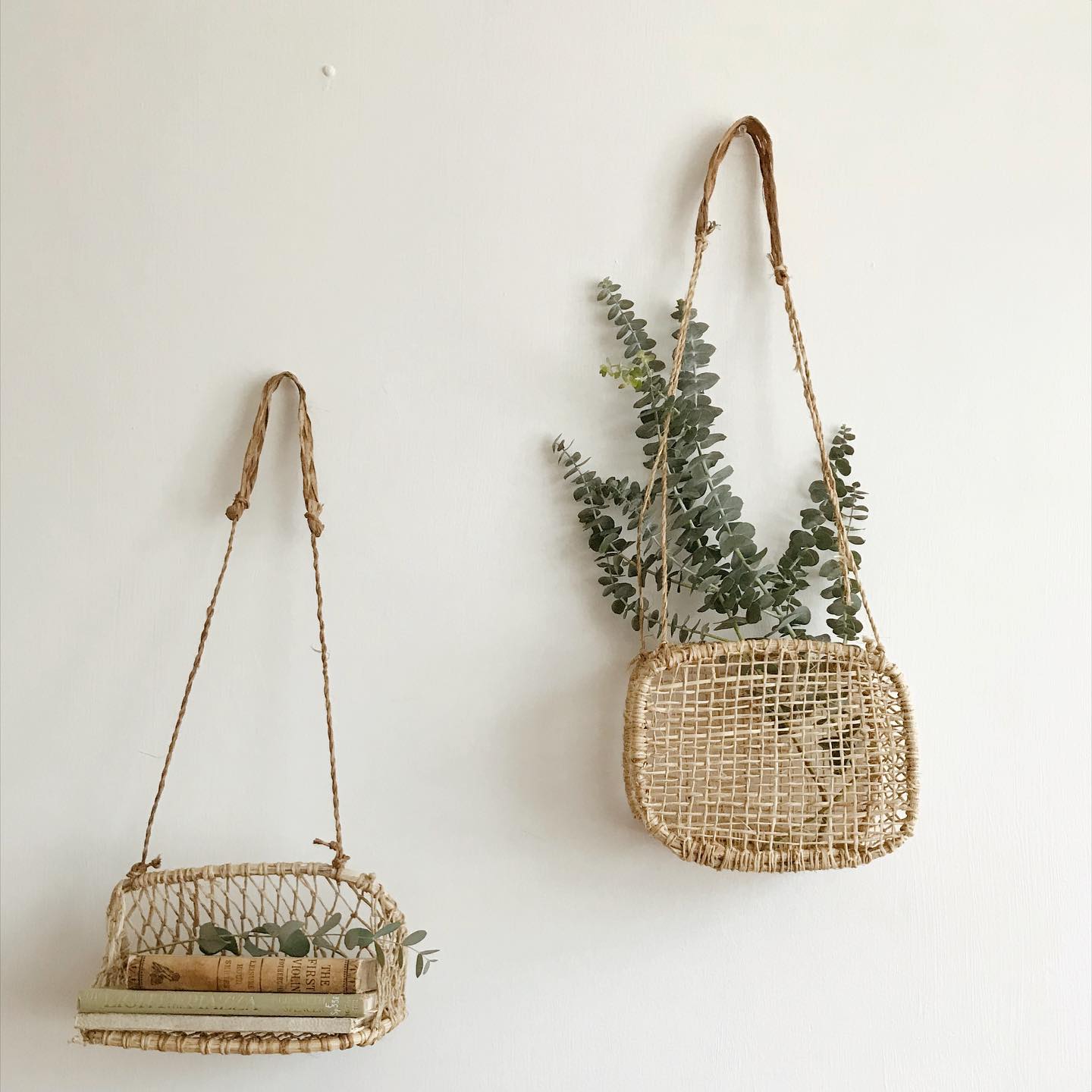 Two jonote woven hanging baskets on a white wall, one filled with eucalyptus branches.