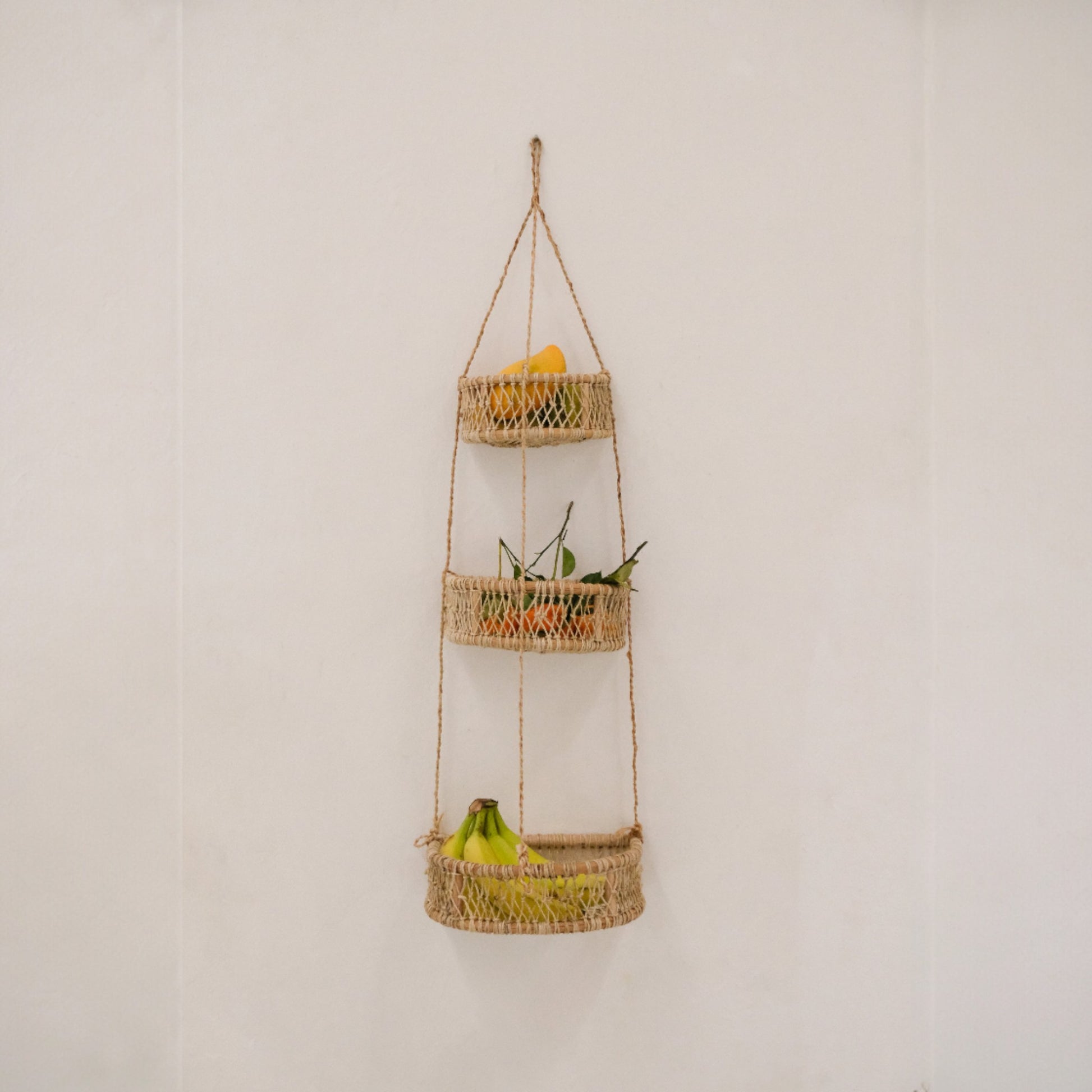 Three-tier woven hanging basket with fruits and plants against a white wall