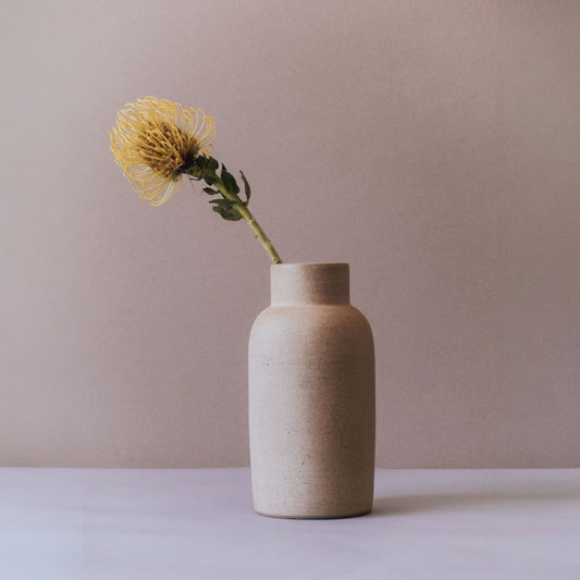 Sand terracotta bottle vase with a single yellow stem on a white surface.