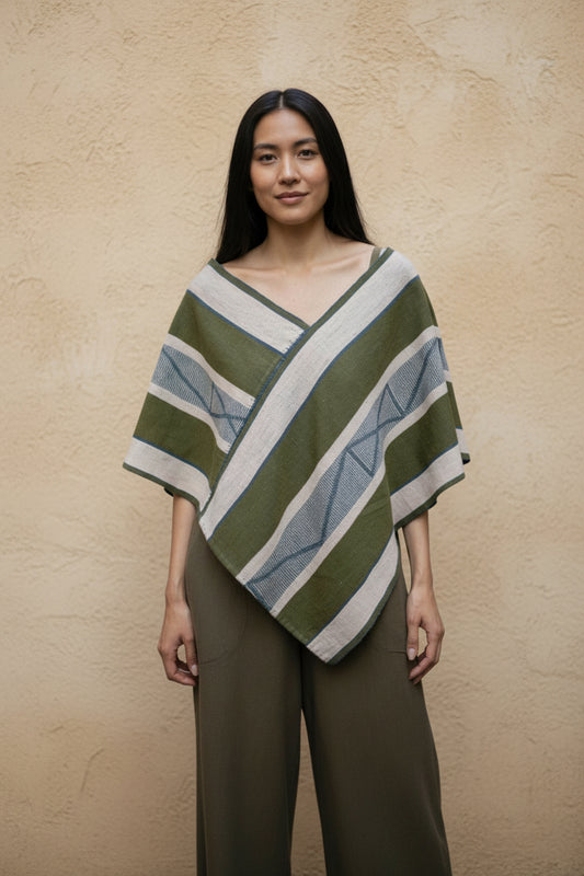 Model wearing a handwoven green and ivory Mexican poncho with geometric pattern, styled over neutral trousers against a warm textured wall