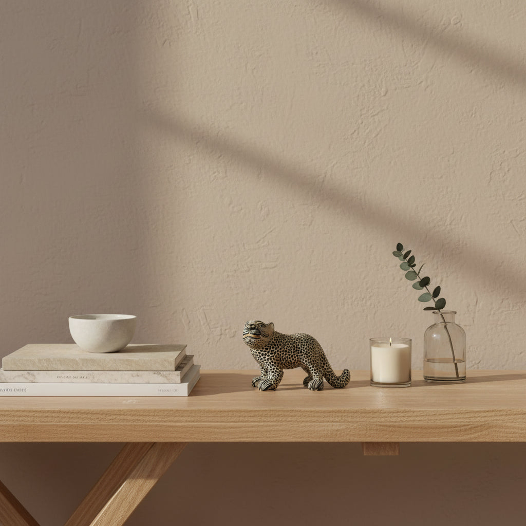 Handmade beige clay jaguar figurine from Amatenango del Valle displayed on a wooden shelf with stacked books, a candle, and a ceramic vase against a beige wall.
