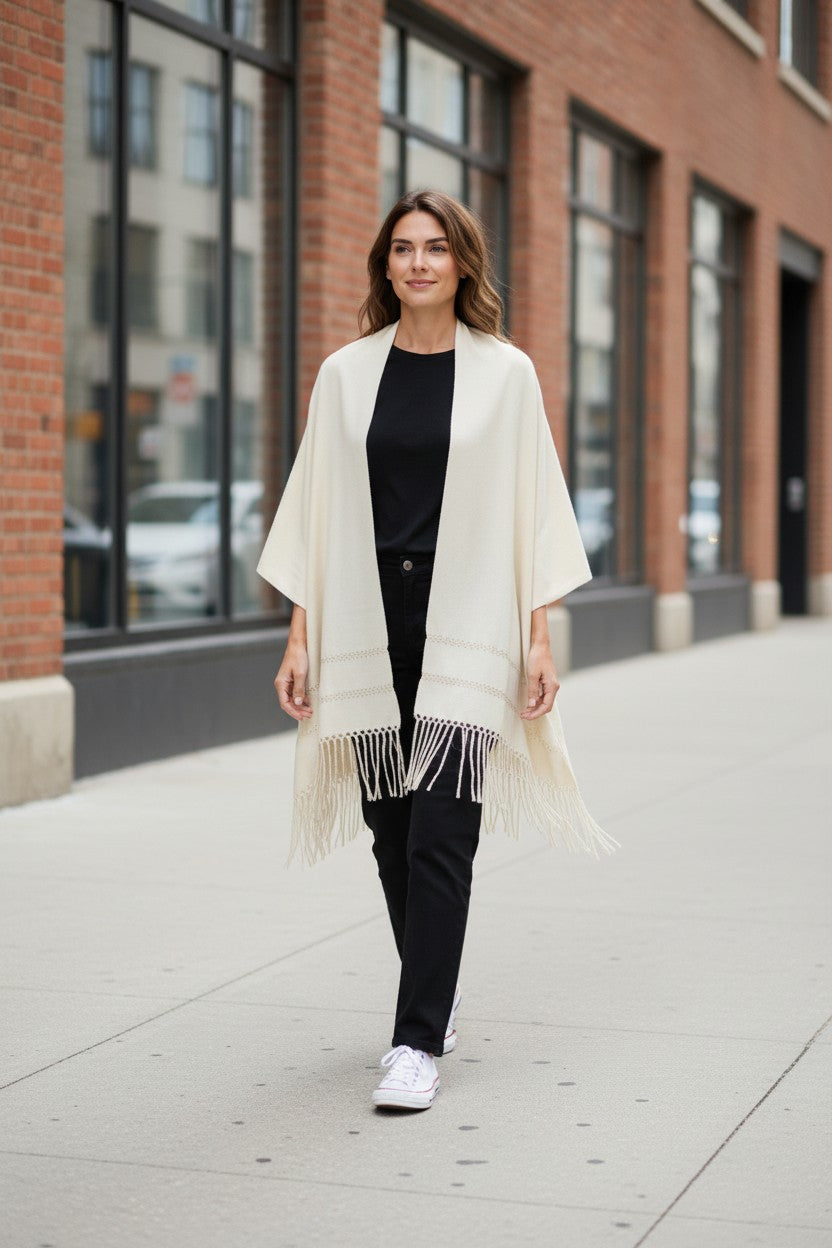 Woman walking on a city sidewalk wearing an ivory handwoven rebozo shawl with fringe, styled casually over a black outfit.